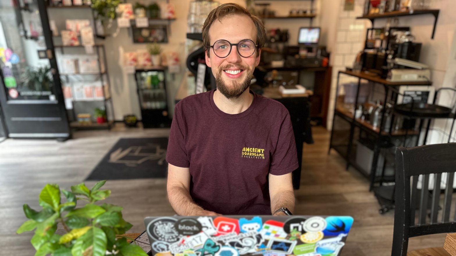 A photo of Klemens Strasser, developer of Ancient Board Game Collection, seated at a coffee shop table behind a laptop that’s covered in various colorful stickers.
