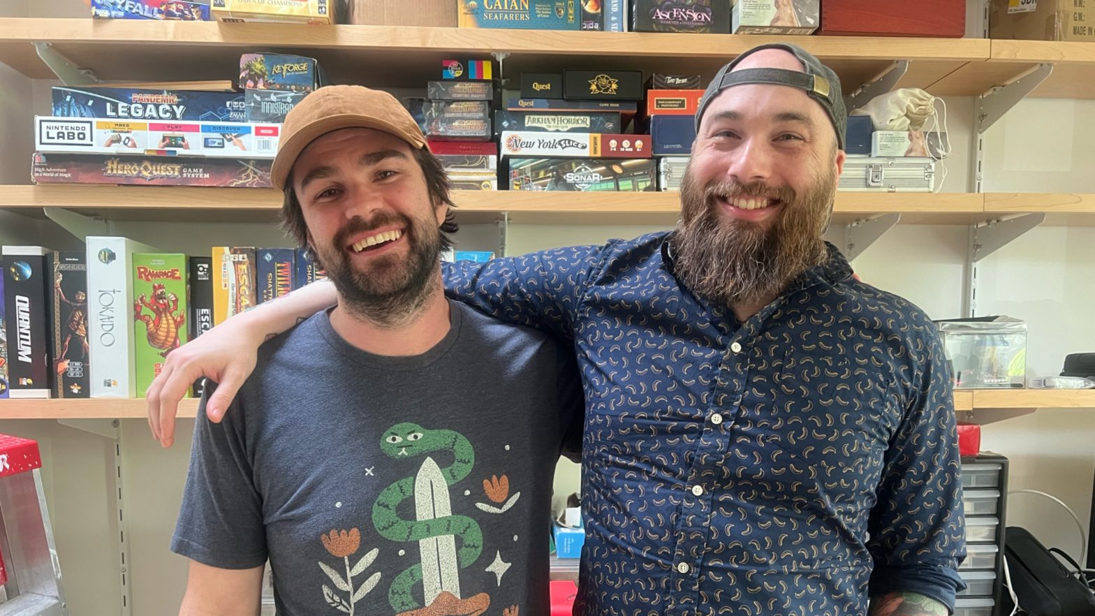Zach Gage, at left, and Jack Schlesinger stand in their studio in front of a series of shelves that hold numerous board games.